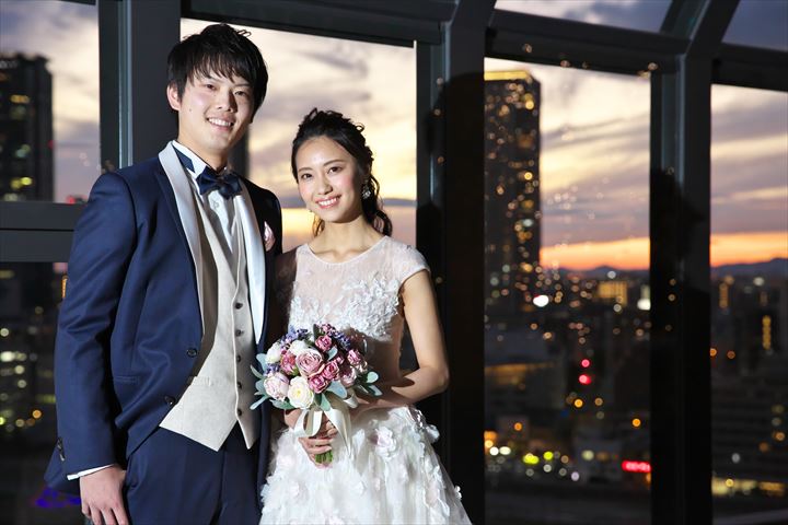 結婚式持ち込みカメラマンが撮影：披露宴お開き後に高層ビルのテラスで夕陽を背にして微笑んでいる新郎新婦